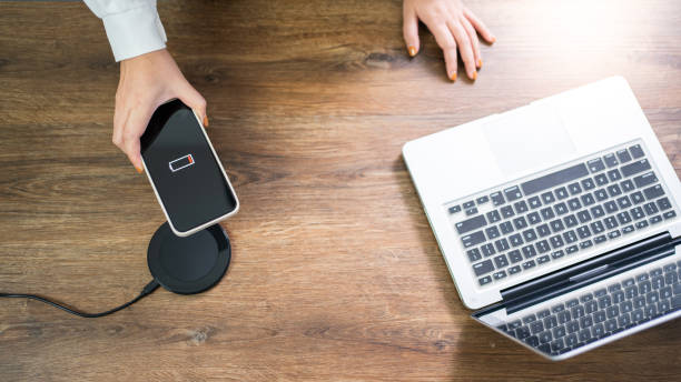 A person places a phone with a low battery on a wireless charging pad next to a laptop on a wooden desk.